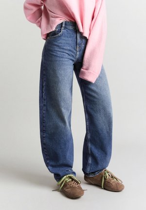 Personne portant un jean bleu ample, un pull rose et des baskets marron avec des lacets verts, debout sur un fond clair.