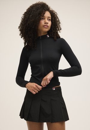 Donna con capelli ricci che indossa una maglia nera a maniche lunghe con zip e una gonna nera plissettata, che sta sistemando la cerniera della maglia.