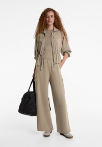 Béžový dvojdielny outfit s presúvacou bundou s vreckami a širokými nohavicami. Modelka nesie čiernu tašku a má na sebe svetlé tenisky.