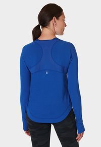 Langarm T-shirt van helderblauwe stof met een textuur op het achterpaneel, afgeronde zoom en drop schouders. Zwarte, gemusterde leggings maken de look compleet.