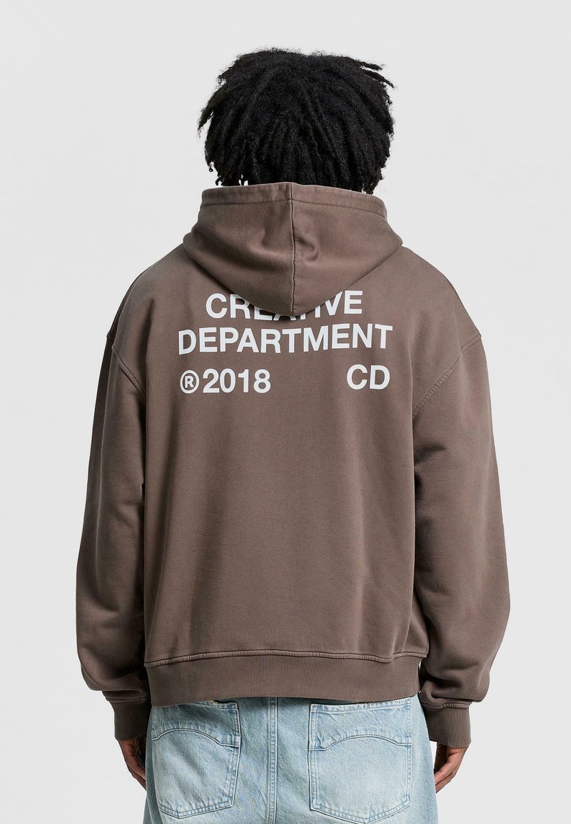 Brown sweatshirt with a hood, featuring a large white text print on the back: "CREATIVE DEPARTMENT 2018 CD." Made of soft fabric, regular fit.