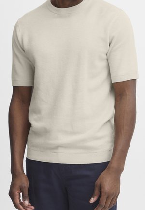 Mann trägt beige kurzärmeliges Strickshirt und dunkelblaue Hose, steht vor schlichtem hellem Hintergrund.