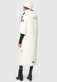 Lang hvid dunjakke med korte ærmer, lynlåslukning og hætte. Bæres over en sort sweater og sorte skinny jeans med ankelstøvler.