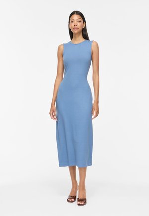 Femme debout portant une robe midi ajustée, sans manches, bleu clair, avec un col rond et des sandales à talons ouvertes marron.