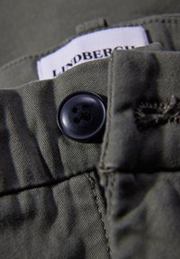 Primer plano de una tela verde oliva que presenta un botón negro y detalles de costura. Etiqueta visible que indica la marca Lindbergh.