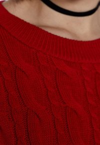Pull en laine rouge avec un motif en torsades, au col large et à la texture travaillée. Porté avec un accessoire de choker noir.