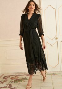 Robe midi noire avec tissu plissé et dentelle au niveau de l'ourlet. Présente un décolleté en V, des manches longues transparentes et une ceinture assortie.