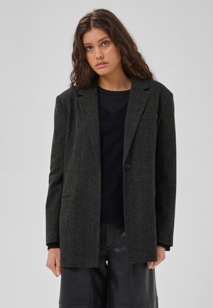 Frau mit langen, welligen Haaren trägt einen dunkelgrauen Blazer, einen schwarzen Pullover und eine schwarze Hose und steht vor einem schlichten, hellen Hintergrund.
