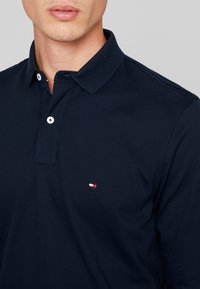 Muž v námořnickém modrém polo tričku s límečkem, dvěma knoflíky a malým červeným, bílým a modrým logem na prsou.