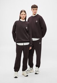 Σκούρα καφέ φούτερ και παντελόνια φόρμας από μαλακό ύφασμα, με διαμάντις γραφικά στο στήθος και στο μηρό, σε συνδυασμό με λευκά πουκάμισα.