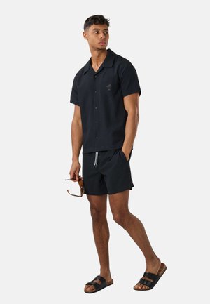 Młody mężczyzna w czarnej koszuli z krótkim rękawem zapinanej na guziki, pasujących szortach, czarnych sandałach, trzyma okulary przeciwsłoneczne, stoi na białym tle.