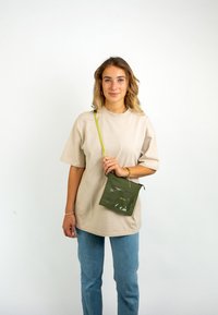 T-shirt oversize beige, cotone leggero, maniche corte. Il modello tiene in mano una pochette verde con pannello frontale trasparente e zip. Jeans blu.