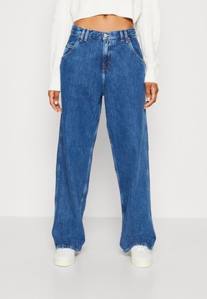 Personne portant un jean bleu large taille haute, un pull court blanc et des baskets blanches, debout devant un fond clair uni.