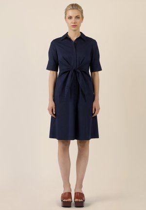 Frau steht frontal und trägt ein marineblaues knielanges Kleid mit halblangen Ärmeln und einer vorderen Bindedetail, kombiniert mit braunen Plateausandalen.