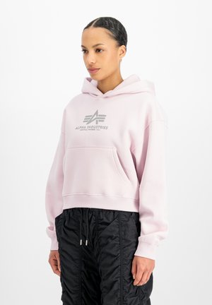 Frau trägt einen hellrosa Alpha Industries Hoodie und schwarze gesteppte Hose und steht vor einem weißen Hintergrund.