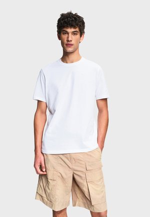 Jeune homme portant un t-shirt blanc uni et un short beige cargo, debout devant un fond gris clair.
