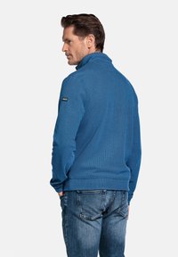 Blauwe, textuurtrui met hoge kraag en lange mouwen. Toon een klein logo op de linker schouder. Gekozen met een versleten spijkerbroek.