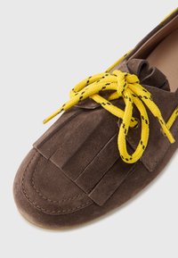 Sapato de camurça castanho com atacadores amarelos e pretos com padrão. Apresenta um detalhe de franjas na parte superior. Biqueira arredondada e costura visível.