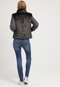 Czarna kamizelka z futra ekologicznego z wysokim kołnierzem, w zestawieniu z szarymi rękawami, noszona na niebieskie dżinsy i srebrne sneakersy. Gładka struktura i prosty design.