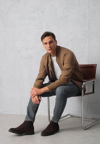 Brązowa kurtka bombowiec na białej koszuli, szare dżinsy i ciemne zamszowe botki Chelsea. Siedzący na metalowym krześle na tle teksturowanego białego tła.