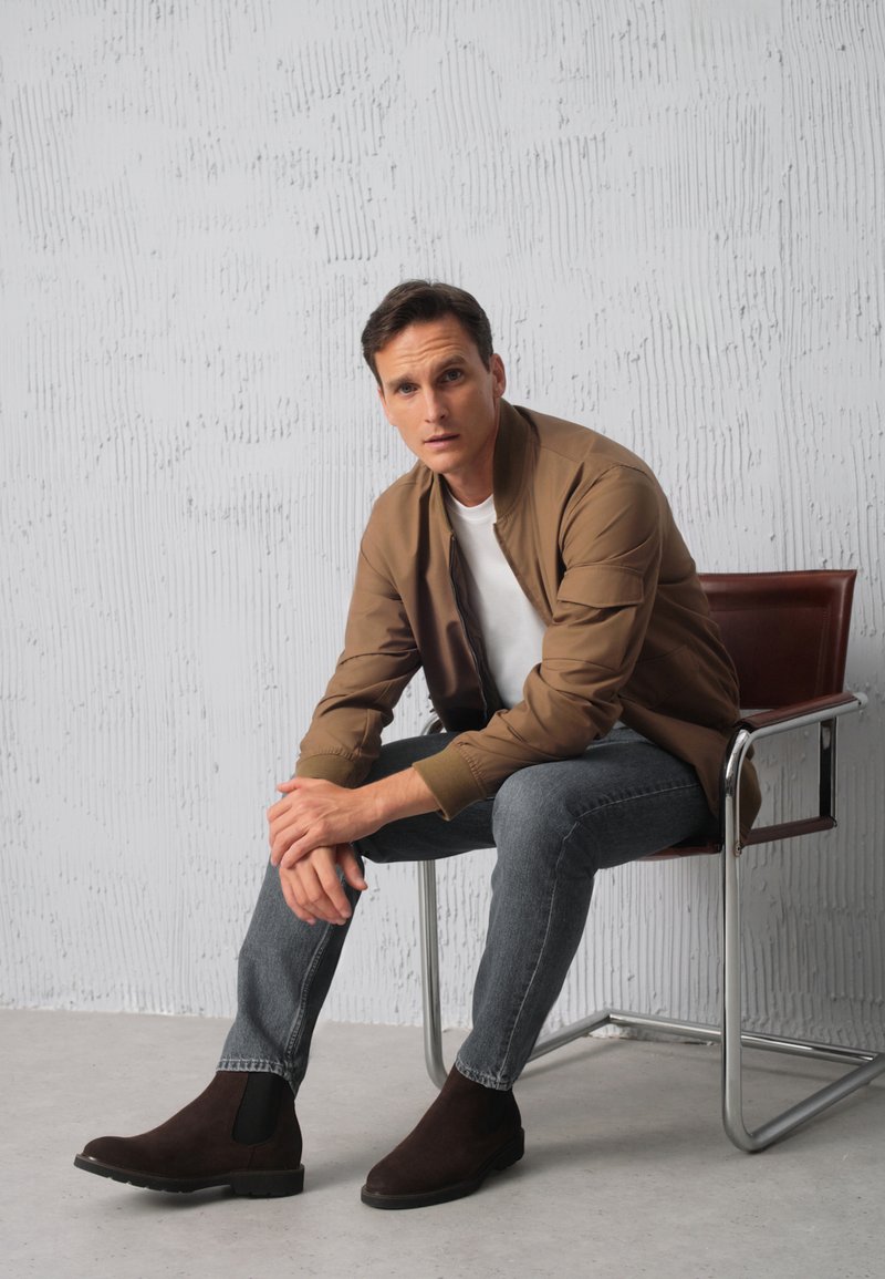 Brązowa kurtka bombowiec na białej koszuli, szare dżinsy i ciemne zamszowe botki Chelsea. Siedzący na metalowym krześle na tle teksturowanego białego tła.