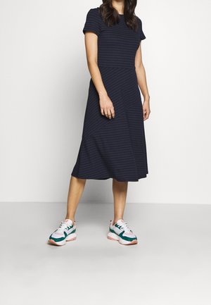 Frau in einem marineblauen Midikleid mit dezenten Streifen und weißen Sneakern mit grünen und pinken Akzenten, stehend auf einem schlichten hellen Boden.