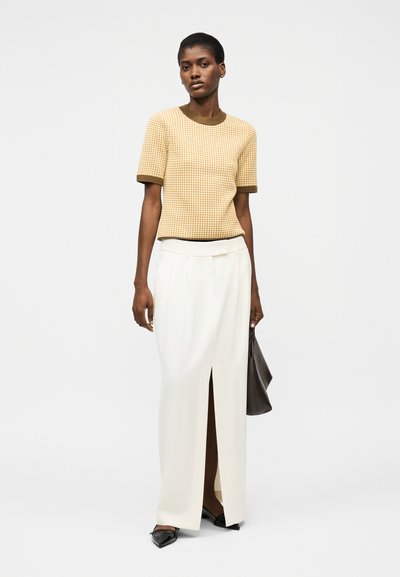 Kvinne iført en kortermet beige og brun rutete topp, hvit høytlivsskjørt med splitt foran, svarte spisse sko, holder en mørk veske.