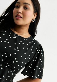 Schwarzes Kleid mit weißen Pünktchen, kurzen Puffärmeln, Raffung an der Taille, begleitet von einer silbernen Halskette und Creolen.