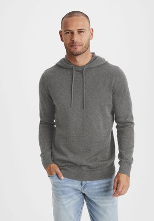 Grauer Kapuzenpullover aus weichem Stoff, mit einer Fronttasche, Kordeln an der Kapuze und gerippten Bündchen, kombiniert mit hellblauen Jeans.