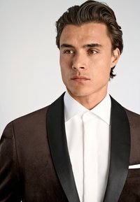 Junger Mann in einem dunkelbraunen Samt-Tuxedojackett mit schwarzen Satin-Revers und einem weißen Hemd, der nach rechts schaut.