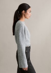 Szary krótki sweter z długimi rękawami, wykonany z miękkiego dzianinowego materiału, noszony z czarnymi spodniami z wysokim stanem. Tło jest neutralne.