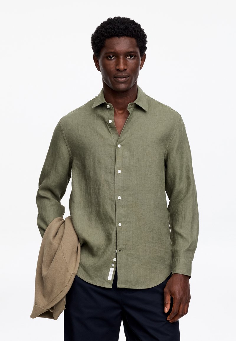 Homme portant une chemise boutonnée olive et tenant une veste beige, debout devant un fond blanc uni.