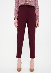 Pantalons ajustés bordeaux avec une coupe slim, longueur cheville, poches slim et accents de boutons dorés à la taille, fabriqués à partir d'un tissu lisse.