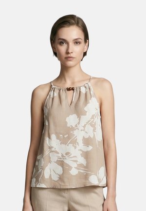 Vrouw met kort haar die een mouwloze beige top draagt met een wit bloemmotief en houten kraaldetail bij de halslijn, staande tegen een effen achtergrond.