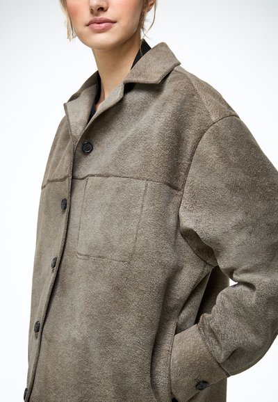 Žena oblečená v šedej semišovej kabáte na gombíky s predným vreckom a golierom, jej ruka vrecku kabáta na bielom pozadí.