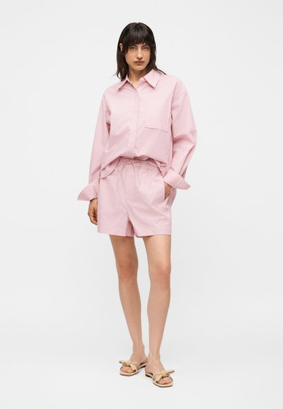 Γυναίκα με ροζ μακρυμάνικο πουκάμισο με τσέπη στο στήθος και ταιριαστά σορτς, στέκεται με τα χέρια στις τσέπες, μπεζ σανδάλια σε λευκό φόντο.
