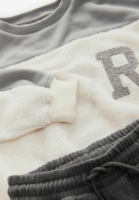 Fleecna dukserica v sivi in kremni barvi z rebrastimi manšetam in strukturiranim detajlom "R". V kompletu s sivimi spodnjimi hlačami na vrvico.