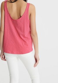 Pink ærmeløs top med lav ryg, lavet af letvægtsstof. Bæres med hvide jeans, der viser en afslappet pasform og glat tekstur.