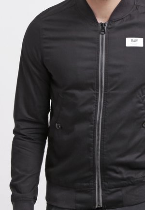 Mand iført en sort bomberjakke med lynlås, knaplommer i siderne og et "RAW"-logo på brystet.