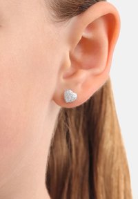 Silvriga hjärtformade örhängen med en strukturerad yta, vilande på örsnibben, som visar upp en kompakt design och en subtil lyster.