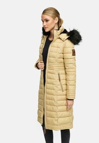 Beige puffjakke med lynlås, ribbet tekstur, lange ærmer og hætte foret med pels. Indeholder en brun accent på den ene ærme.