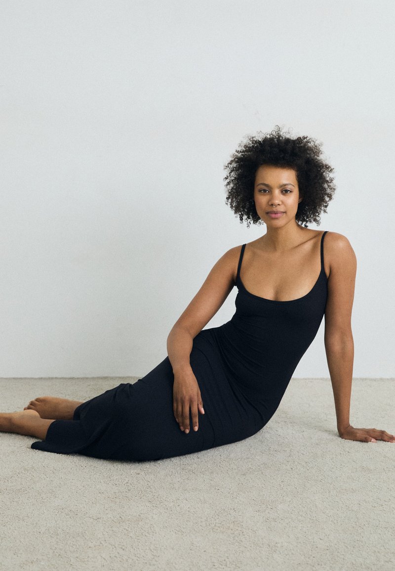 Robe maxi noire sans manches avec un corps ajusté, présentant un décolleté rond et une fente à l'ourlet, stylée dans une pose assise détendue.