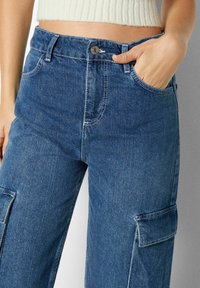 Denim cargobyxor i mörkblått med hög midja, stora sidofickor och kontrasterande sömnadsdetaljer. Mjuk textur och avslappnad passform.