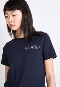 T-shirt en coton bleu marine avec un logo imprimé blanc et du texte. Design à manches courtes et col rond, texture lisse. Style minimaliste.