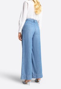 Mujer con cabello largo y rubio, vestida con pantalones vaqueros de pierna ancha de color azul claro, camisa blanca de manga larga y zapatos de tacón bajo de color gris claro, de pie contra un fondo blanco.