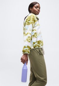 Νέα γυναίκα με πράσινο και λευκό fleece με σχέδια και πράσινο παντελόνι, κρατώντας μια μωβ παγούρι νερού πάνω σε απλό φόντο.