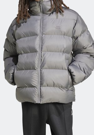 Personne portant une doudoune grise adidas avec les mains dans les poches et un pantalon noir adidas sur un fond uni.