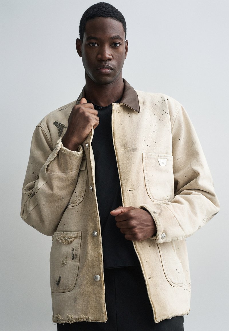 Jeune homme portant une veste beige usée avec un col marron par-dessus une chemise noire, tenant les revers de la veste contre un fond gris uni.