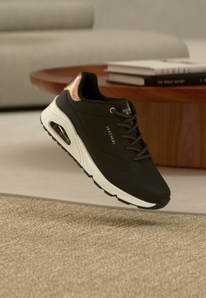 Black athletic shoe with a textured upper, gold accent at the heel, white midsole, and black rubber sole. Minimal logo detail on the side.