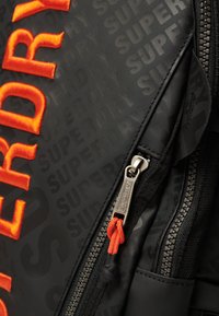 Czarny plecak z wytłoczonym pomarańczowym haftowanym logo "SUPERDRY", teksturowana powierzchnia z powtarzającym się brandingiem oraz srebrny zamek błyskawiczny z pomarańczowym uchwytem.